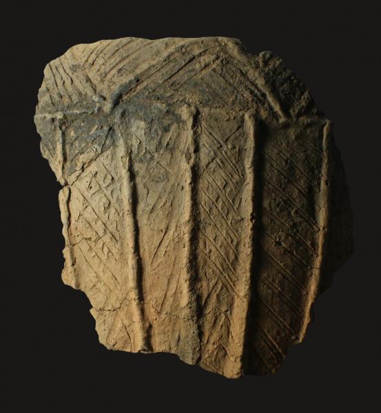 A photograph of a large fragment of pottery decorated with vertical grooves and incised with horizontal lines on the body and v shaped lines around the rim